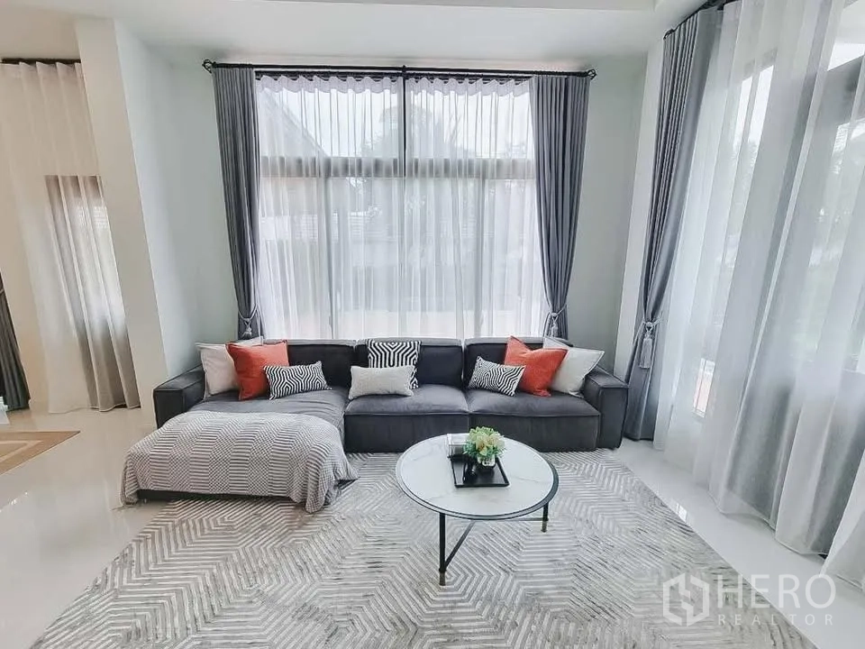 Detached House for rent or sale in Bang Phli, Samut Prakan - Light-filled sitting area with L-shaped sofa, patterned rug, and floor-to-ceiling windows.