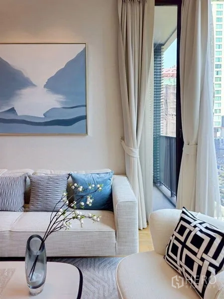 Condominium for rent or sale in Pathum Wan, Bangkok - Sunlit living room with light sofa, abstract artwork, and floor-to-ceiling curtains opening to a balcony.