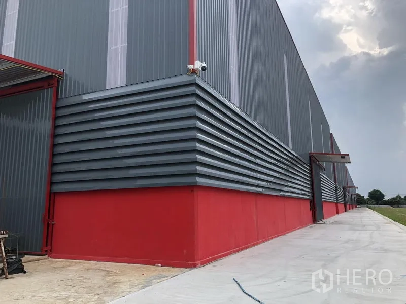 Warehouse for rent in Khlong Luang, Pathum Thani - Side corner of warehouse with corrugated metal cladding, red base, CCTV camera and covered entry.