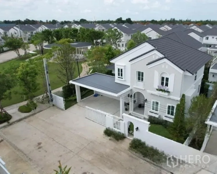 独立式住宅 出租在Bang Phli, Samut Prakan - 鸟瞰两层白色独栋别墅，带电动门庭院、车棚与园景公园的安保社区。