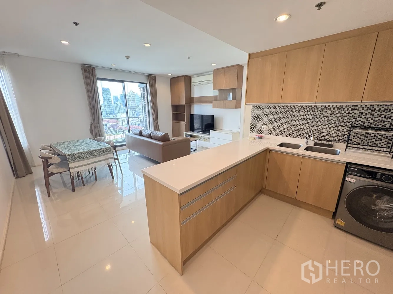 Condominium for rent in Ratchathewi, Bangkok - Open-plan living and dining area with L-shaped kitchen, mosaic backsplash, washing machine, sofa, dining table, and city view through large windows.