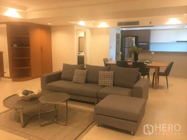 Condominium for rent in Watthana, Bangkok - Open-plan living room with grey sectional sofa, nesting coffee tables, and a dining area beside a modern kitchen.