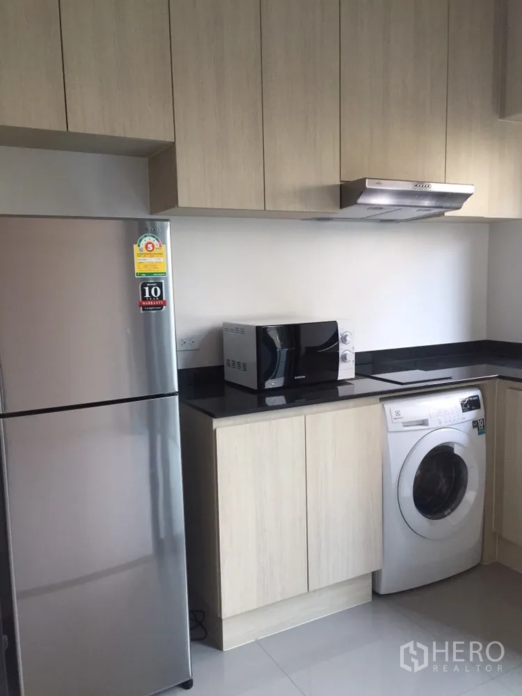 Townhouse for rent in Suan Luang, Bangkok - Modern kitchen with light wood cabinets, black countertops, refrigerator, microwave, and washing machine.