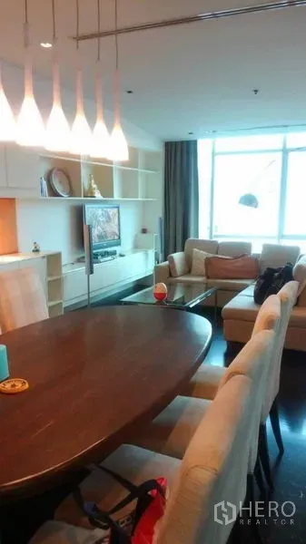 Condominium for sale in Bang Rak, Bangkok - View across dining and living space with pendant lights, shelving and sofa beside a large window.