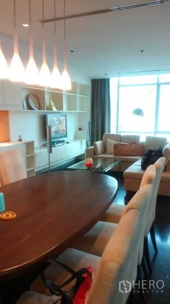 Condominium for sale in Bang Rak, Bangkok - View across dining and living space with pendant lights, shelving and sofa beside a large window.