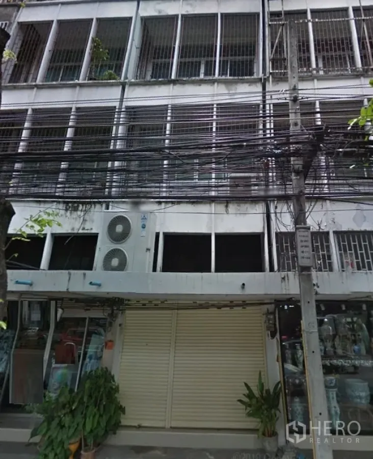 店屋 出租在Phra Khanong, Bangkok - 多层店屋外立面，可见金属格栅窗、关闭的卷闸门、玻璃门面、空调外机与密集的街边电缆。