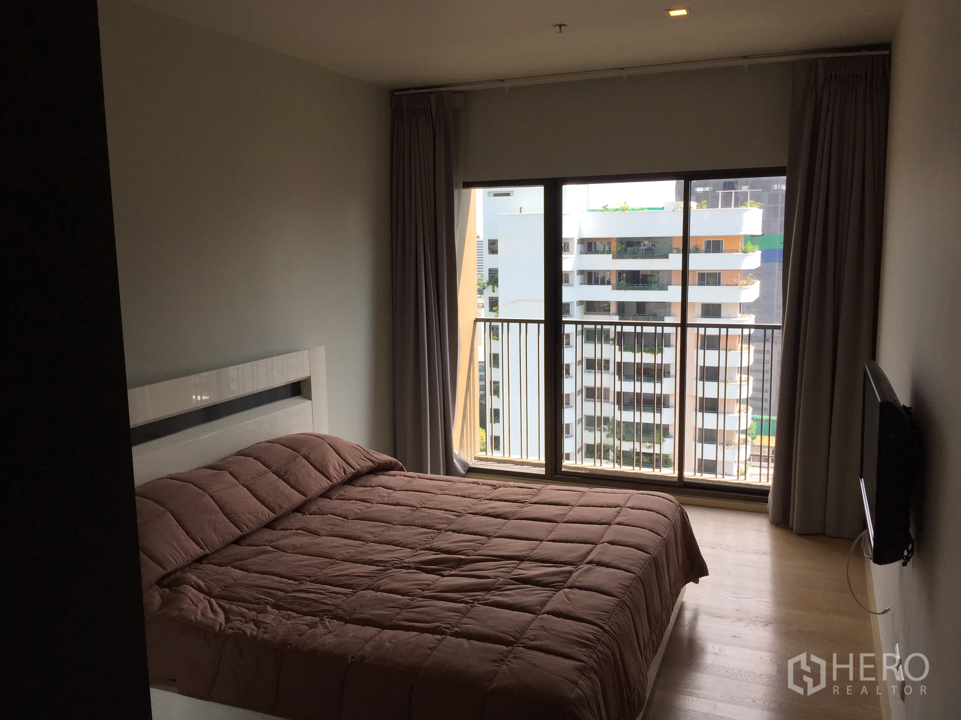 คอนโด ให้เช่าในBangkok - ห้องนอนสว่างพร้อมหน้าต่างสูง ระเบียงวิวเมือง พื้นไม้ และผ้านวมโทนอุ่น.