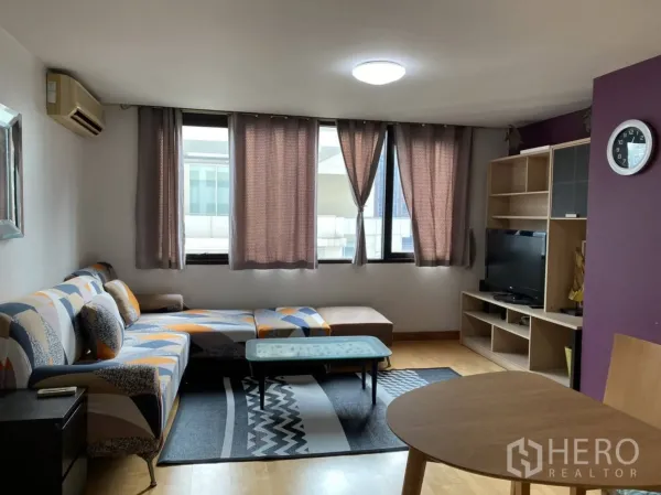 คอนโด ให้เช่าในPathum Wan, Bangkok - มุมนั่งเล่นมีหน้าต่างบานใหญ่ โซฟาลวดลาย ชั้นวางทีวี และผนังสีม่วง