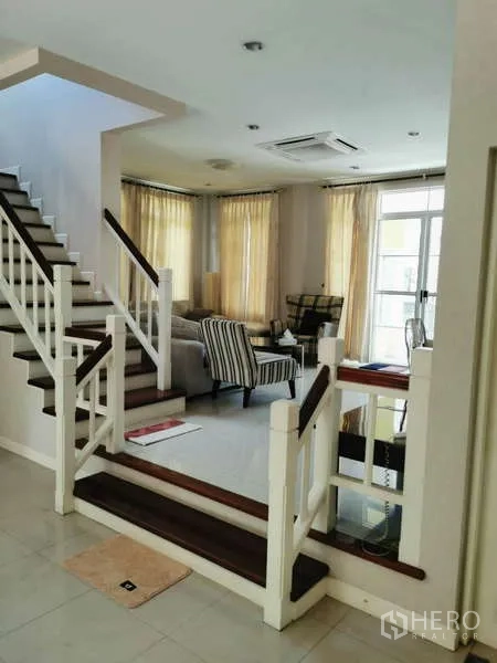 บ้าน ให้เช่า - พื้นที่นั่งเล่นแบบโอเพ่นพร้อมบันไดสีขาว เก้าอี้นวม และหน้าต่างบานใหญ่ผ้าม่านสีครีม