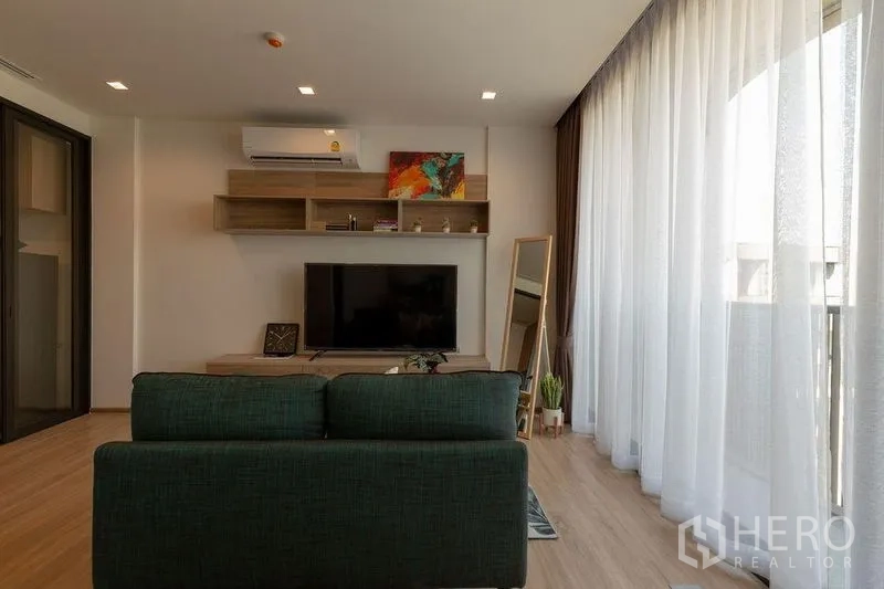 คอนโด ให้เช่าในWatthana, Bangkok - ห้องนั่งเล่นโซฟาสีเขียวหันเข้าหาทีวี ผ้าม่านโปร่งเต็มผนังเปิดสู่ระเบียง