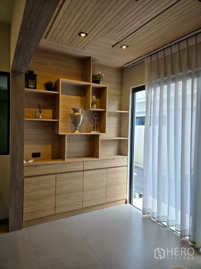 Townhouse for rent in Bang Phli, Samut Prakan - Built-in wooden display shelves and cabinet next to a full-height window with sheer curtains.