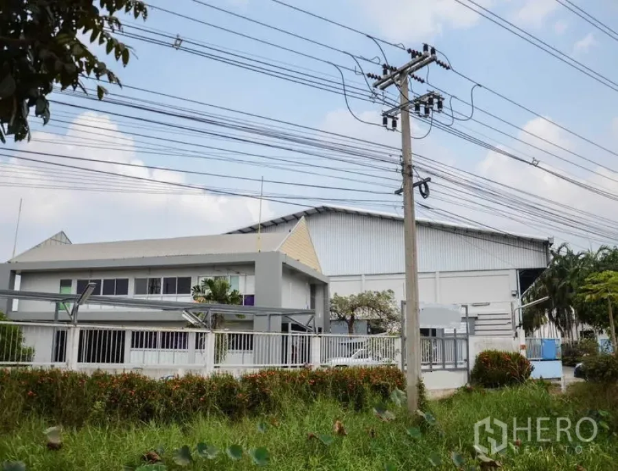 Factory for rent in Pathum Thani - Gated factory complex with office building and large warehouse seen from the road with overhead power lines.
