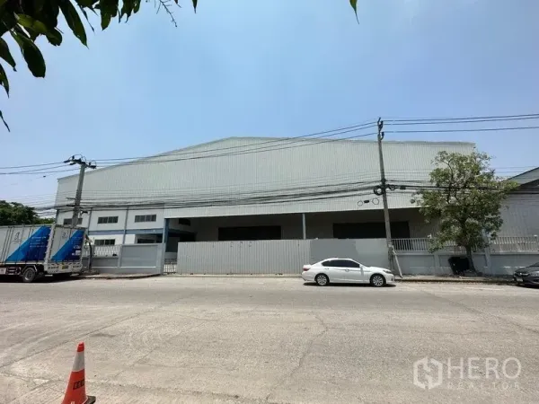 Warehouse for rent in Bang Phli, Samut Prakan - Street view of a large metal-clad warehouse with gated frontage, power lines, and parked cars under a clear sky.