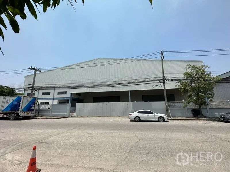 Warehouse for rent in Bang Phli, Samut Prakan - Street view of a large metal-clad warehouse with gated frontage, power lines, and parked cars under a clear sky.