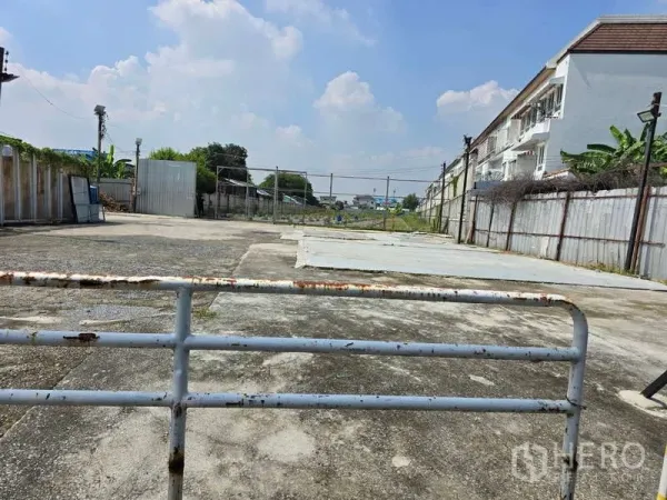 ที่ดิน ให้เช่าในPrawet, Bangkok - ภาพมุมต่ำของที่ดินพื้นคอนกรีตกว้าง มีกำแพงรั้วและประตูเหล็ก ข้างเคียงเป็นทาวน์โฮม ใต้ท้องฟ้าแจ่มใส