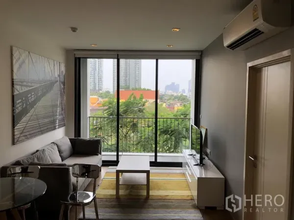 Condominium for rent in Watthana, Bangkok - Bright living room with floor-to-ceiling windows opening to a balcony with leafy city views.