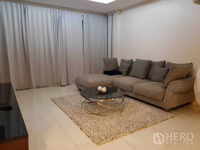 Detached House for rent in Bang Phli, Samut Prakan - Cozy lounge with a gray L-shaped sofa, round glass coffee table, rug and full-length curtains.