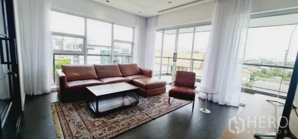 公寓 出租在Watthana, Bangkok - 转角客厅，落地窗、皮沙发、花纹地毯与城市景观。