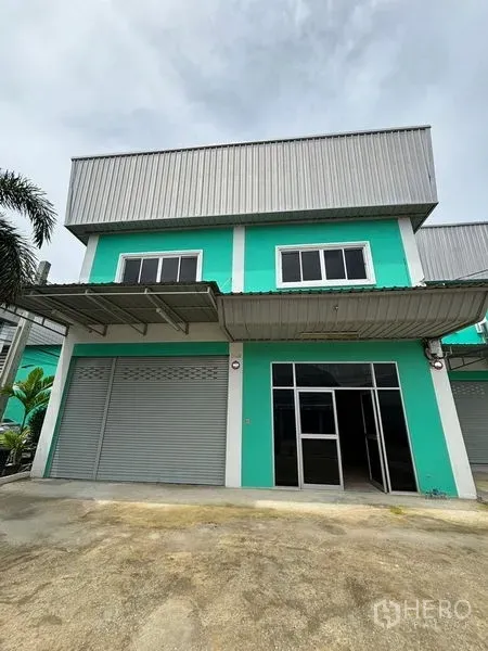 Warehouse for rent in Phra Samut Chedi, Samut Prakan - Two-storey teal warehouse exterior with roller shutters, glass entry and covered loading awning in Phra Samut Chedi.