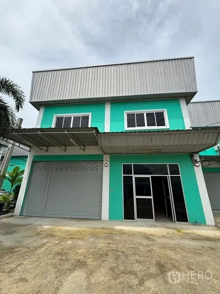 Warehouse for rent in Phra Samut Chedi, Samut Prakan - Two-storey teal warehouse exterior with roller shutters, glass entry and covered loading awning in Phra Samut Chedi.