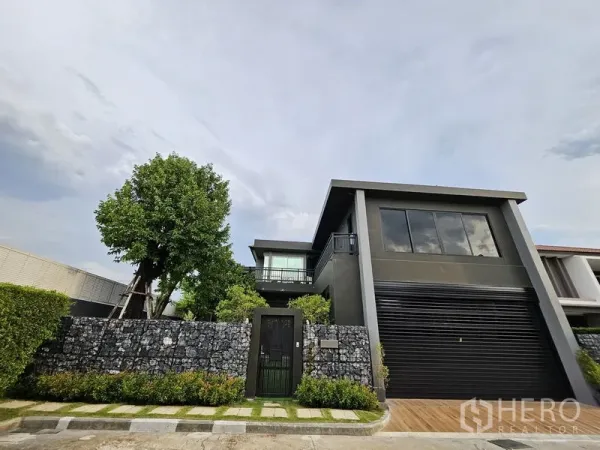 Detached House for sale in Thawi Watthana, Bangkok - Wider front view of the contemporary home behind a gabion stone fence and a mature tree.