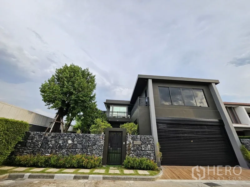 Detached House for sale in Thawi Watthana, Bangkok - Wider front view of the contemporary home behind a gabion stone fence and a mature tree.