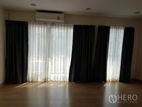 联排别墅 出租在Bang Na, Bangkok - 采光充足的客厅，落地窗搭配纱帘与遮光窗帘，木地板的邦纳联排别墅。