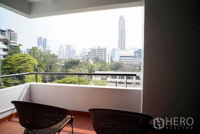 Condominium for rent in Watthana, Bangkok - Covered balcony with two rattan chairs overlooking Bangkok skyline and greenery.