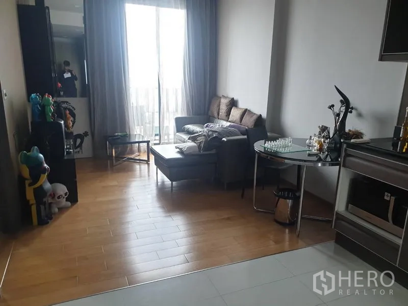 公寓 出租在Khlong Toei, Bangkok - 明亮客厅，灰色转角沙发与脚凳、木地板，以及通向阳台的推拉玻璃门与薄纱窗帘。