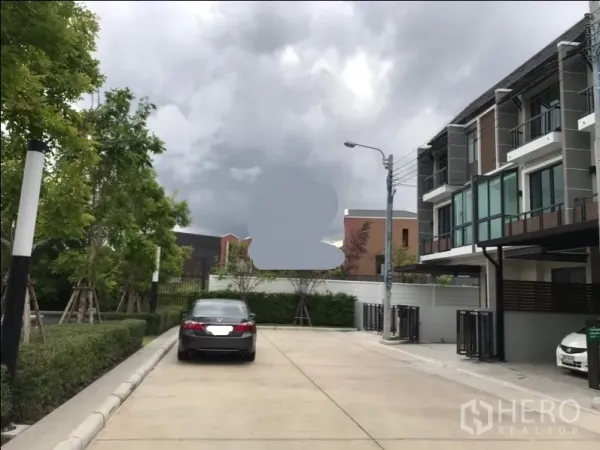 联排别墅 出售在Lat Phrao, Bangkok - 曼谷Lat Phrao现代三层联排别墅外观，社区内道路宽阔，带门前停车位与绿化，在多云天空下。