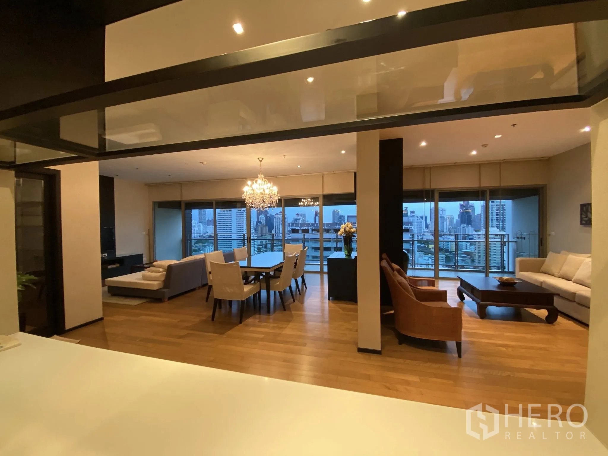 Condominium for rent in Watthana, Bangkok - Spacious lounge and dining with floor-to-ceiling windows, balcony, and city skyline views at dusk.