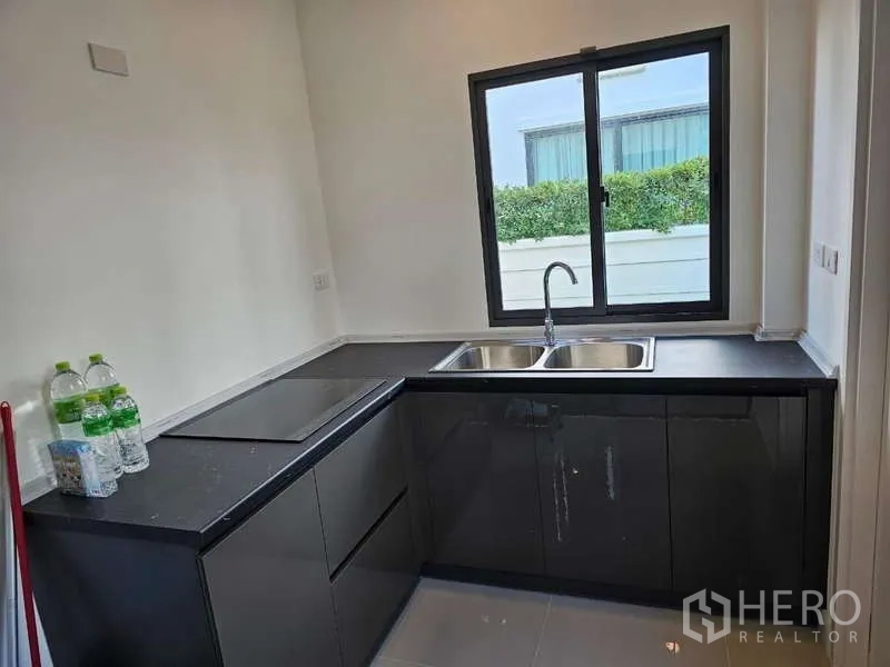 Detached House for rent in Bang Phli, Samut Prakan - L-shaped kitchen with black cabinets, electric cooktop and double sink by a window.