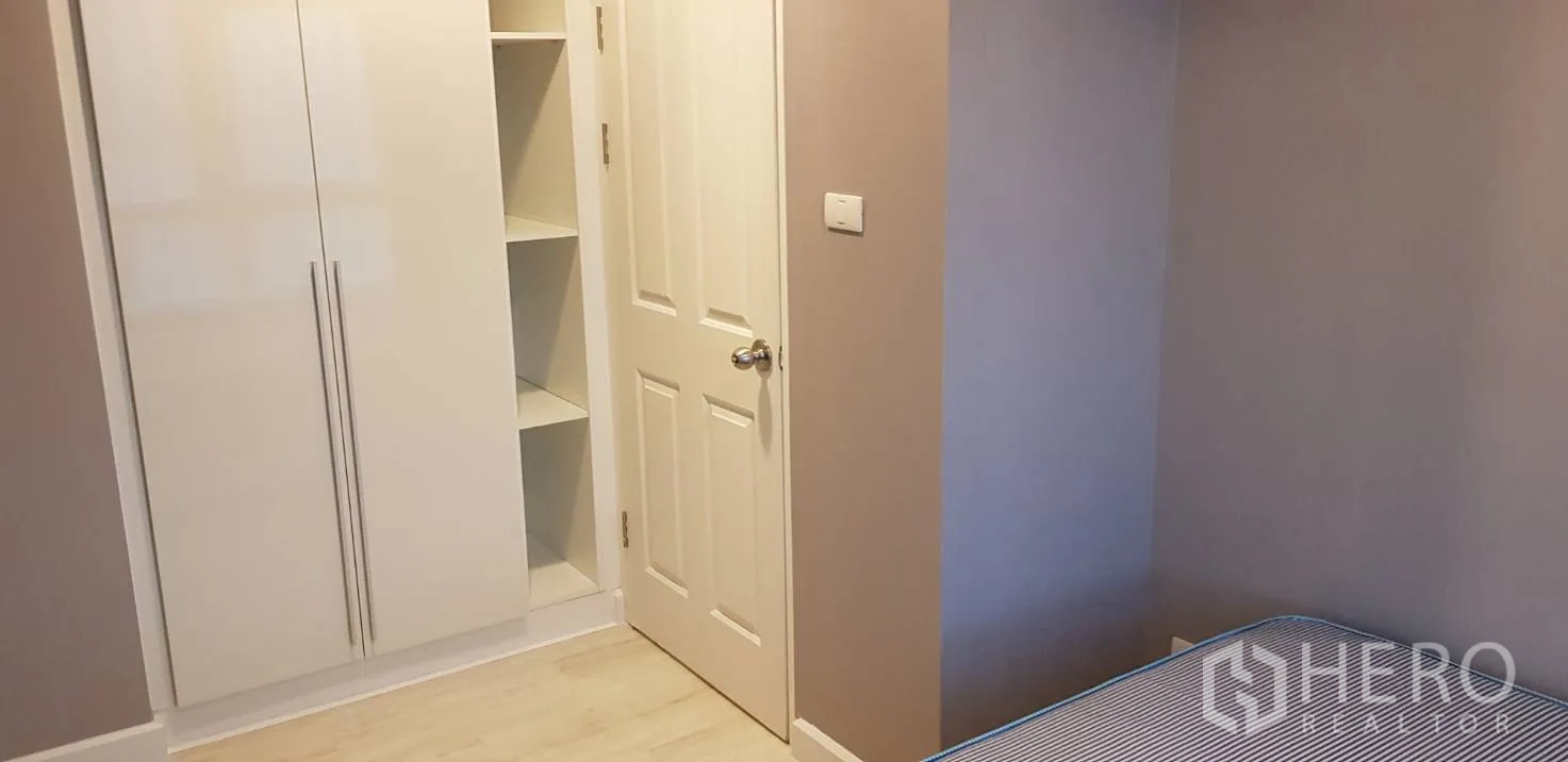 Condominium for rent in Huai Khwang, Bangkok - Bedroom with built-in white wardrobe, open shelving and light laminate floors.