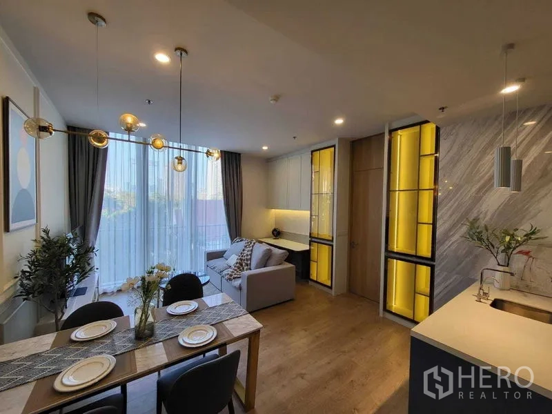 Condominium for rent in Watthana, Bangkok - Dining table and lounge facing floor‑to‑ceiling sheer curtains and balcony, with illuminated built‑in cabinets and a marble kitchen wall.