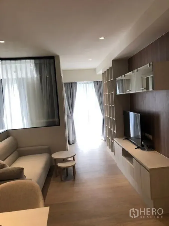 Condominium for rent in Watthana, Bangkok - Cozy living area with sofa, TV wall unit, shelving and floor-to-ceiling curtained window.