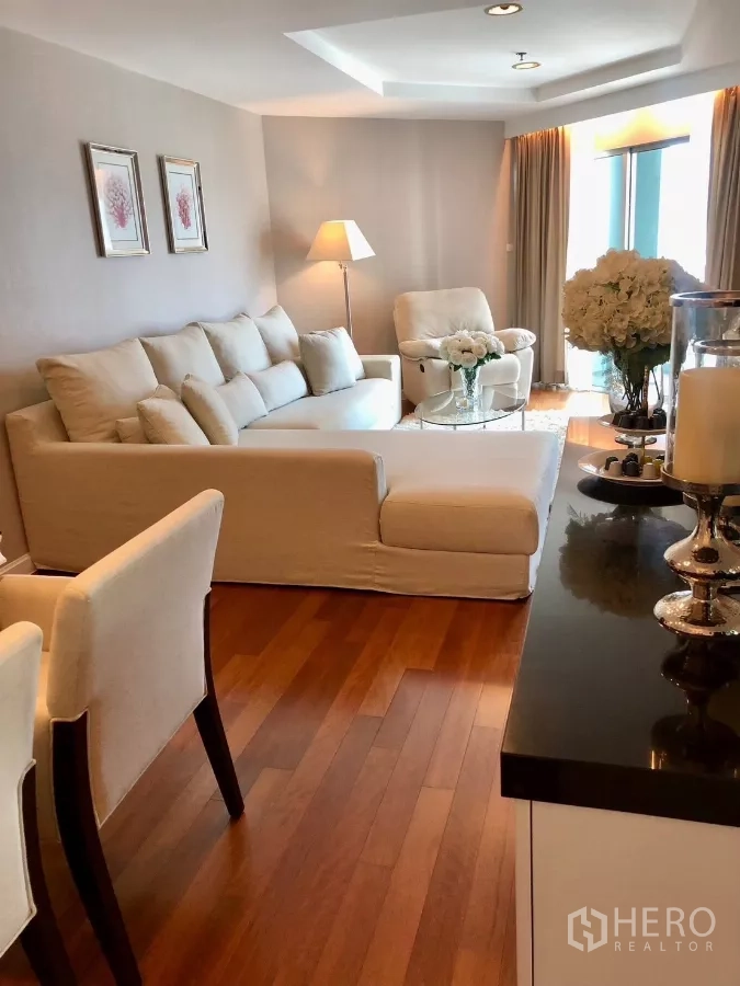 Condominium for rent in Huai Khwang, Bangkok - Cozy lounge with L‑shaped cream sofa, armchair and warm wood floors beside full‑height curtains.