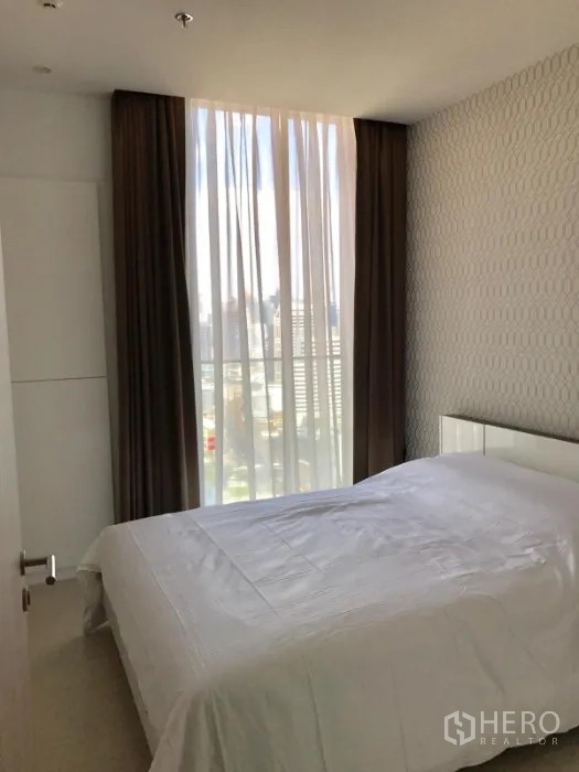 Condominium for rent or sale in Pathum Wan, Bangkok - Cozy bedroom with patterned wallpaper, sheer and blackout curtains, and a city view.