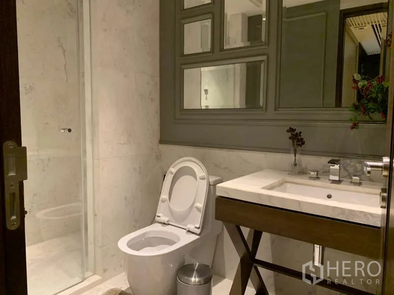 Condominium for rent in Khlong Toei, Bangkok - Compact bathroom with marble walls, glass shower enclosure and sleek vanity.