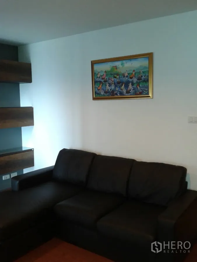 Condominium for rent in Huai Khwang, Bangkok - Living room with black L-shaped leather sofa, framed artwork, floating shelves and wood flooring.