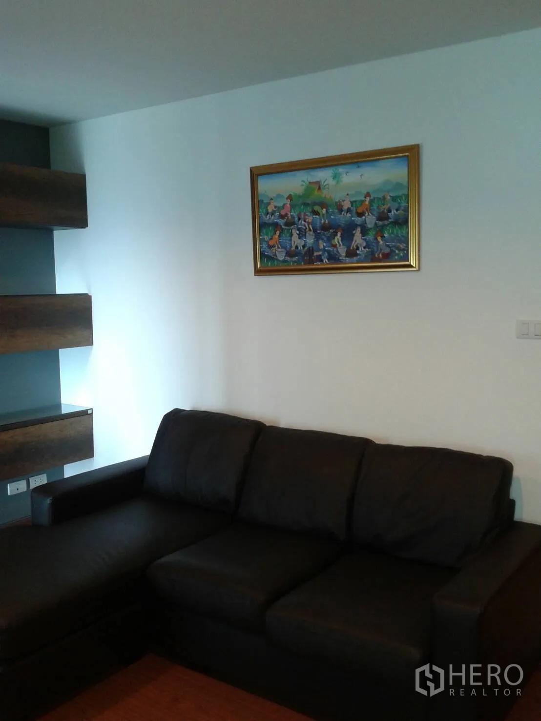 Condominium for rent in Huai Khwang, Bangkok - Living room with black L-shaped leather sofa, framed artwork, floating shelves and wood flooring.