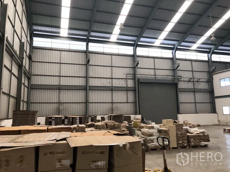 Factory for rent in Khlong Luang, Pathum Thani - Wide warehouse bay with skylights, steel beams, stored pallets and a large roller shutter door.