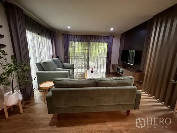 独立式住宅 出租在Prawet, Bangkok - 客厅配绿色沙发、电视墙与落地窗帘，氛围温馨。