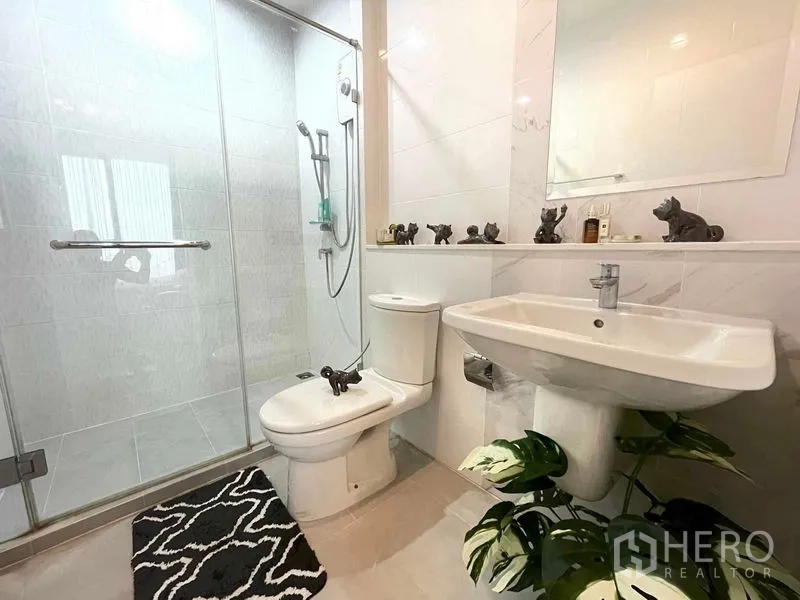 Condominium for sale in Huai Khwang, Bangkok - Modern bathroom with glass-enclosed shower, white tiles, pedestal sink and toilet, accented by a small plant.