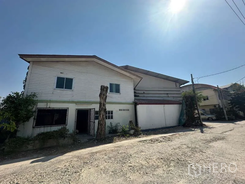 Warehouse for rent in Suan Luang, Bangkok - Street view of a warehouse in Suan Luang with a two‑storey office wing and wide gate under bright sun.