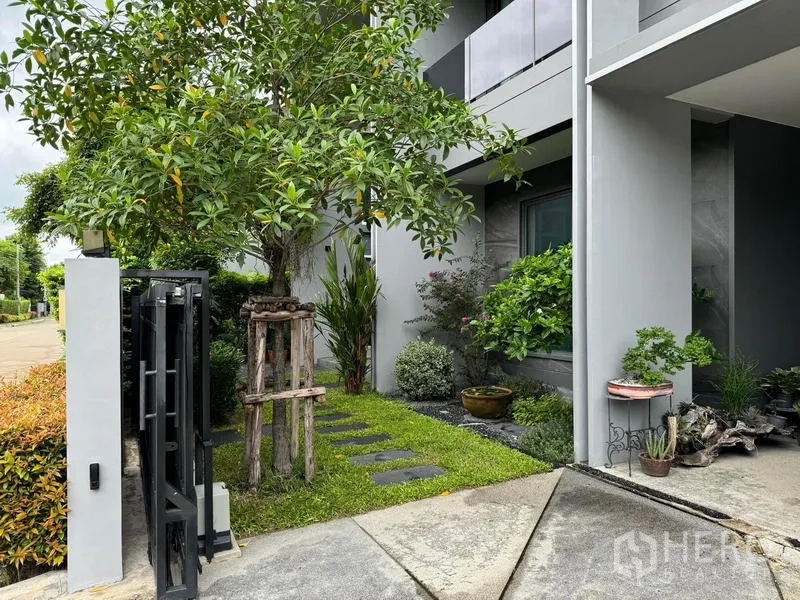 Detached House for rent in Bang Kapi, Bangkok - Front yard view showing gate, mature tree and stepping‑stone path beside the covered carport.