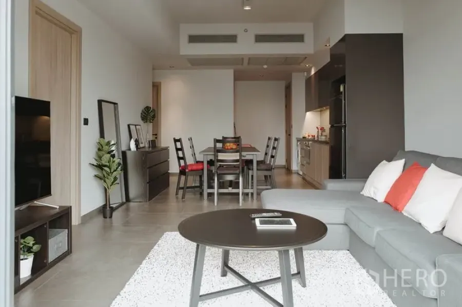 Condominium for rent in Watthana, Bangkok - Open-plan living and dining area with grey sectional sofa, round coffee table, TV console, and a modern kitchen in a Sukhumvit condo.