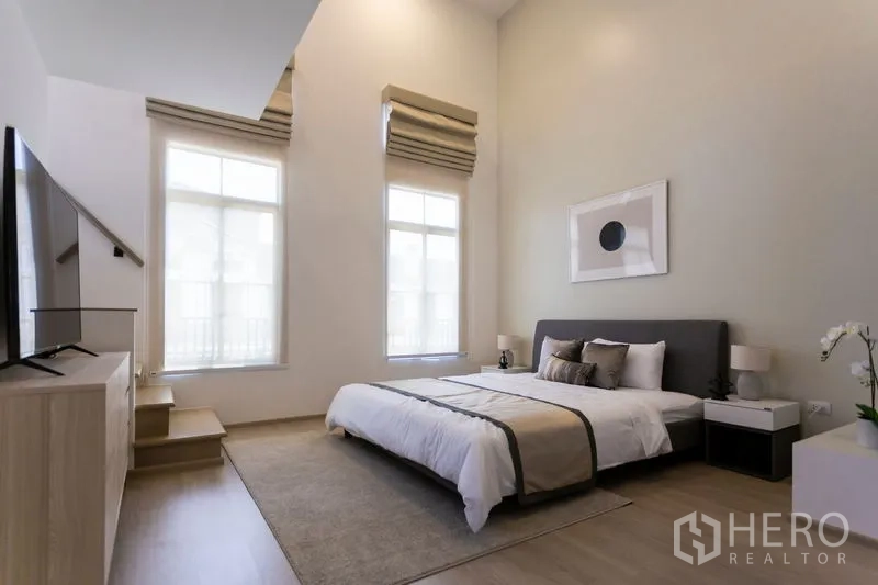 Townhouse for rent in Bang Phli, Samut Prakan - Master bedroom with high ceiling, tall windows, upholstered bed and large TV.