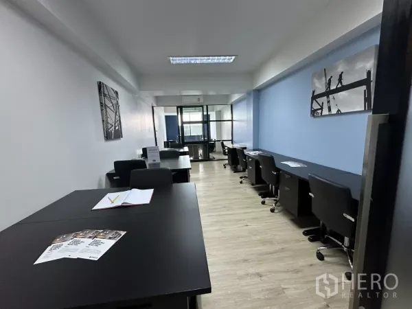 Office Space for rent in Watthana, Bangkok - Open-plan office with rows of black desks, blue accent wall, light wood flooring, and a glass-partitioned meeting room at the end.
