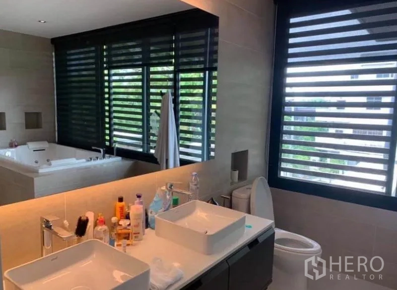 Townhouse for rent in Din Daeng, Bangkok - Master bathroom with double vanity, large mirror and windows with black blinds, jacuzzi in background.