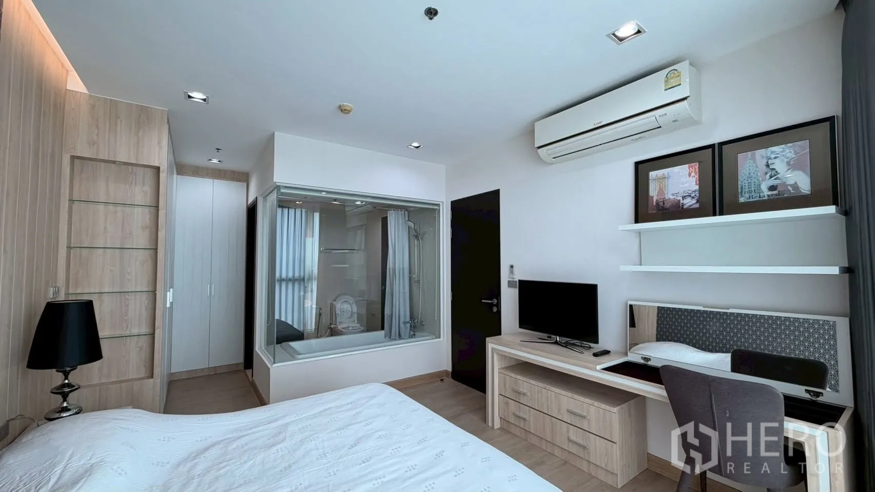 Condominium for rent or sale in Watthana, Bangkok - Bedroom looking toward a glass-partitioned bathroom with bathtub and shower, wardrobe, and work desk with TV.